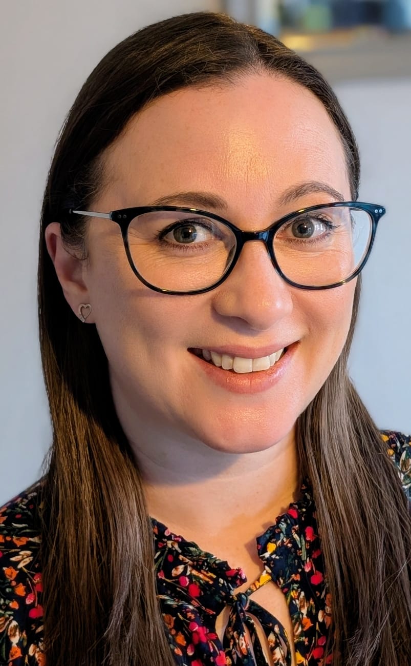 Beth Richard is a woman with long brown hair, wearing glasses and a multicoloured dress.