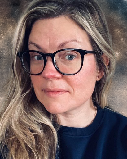 Woman with long hair swept over one shoulder, her head is turned to the side.  She is wearing black rimmed glasses.