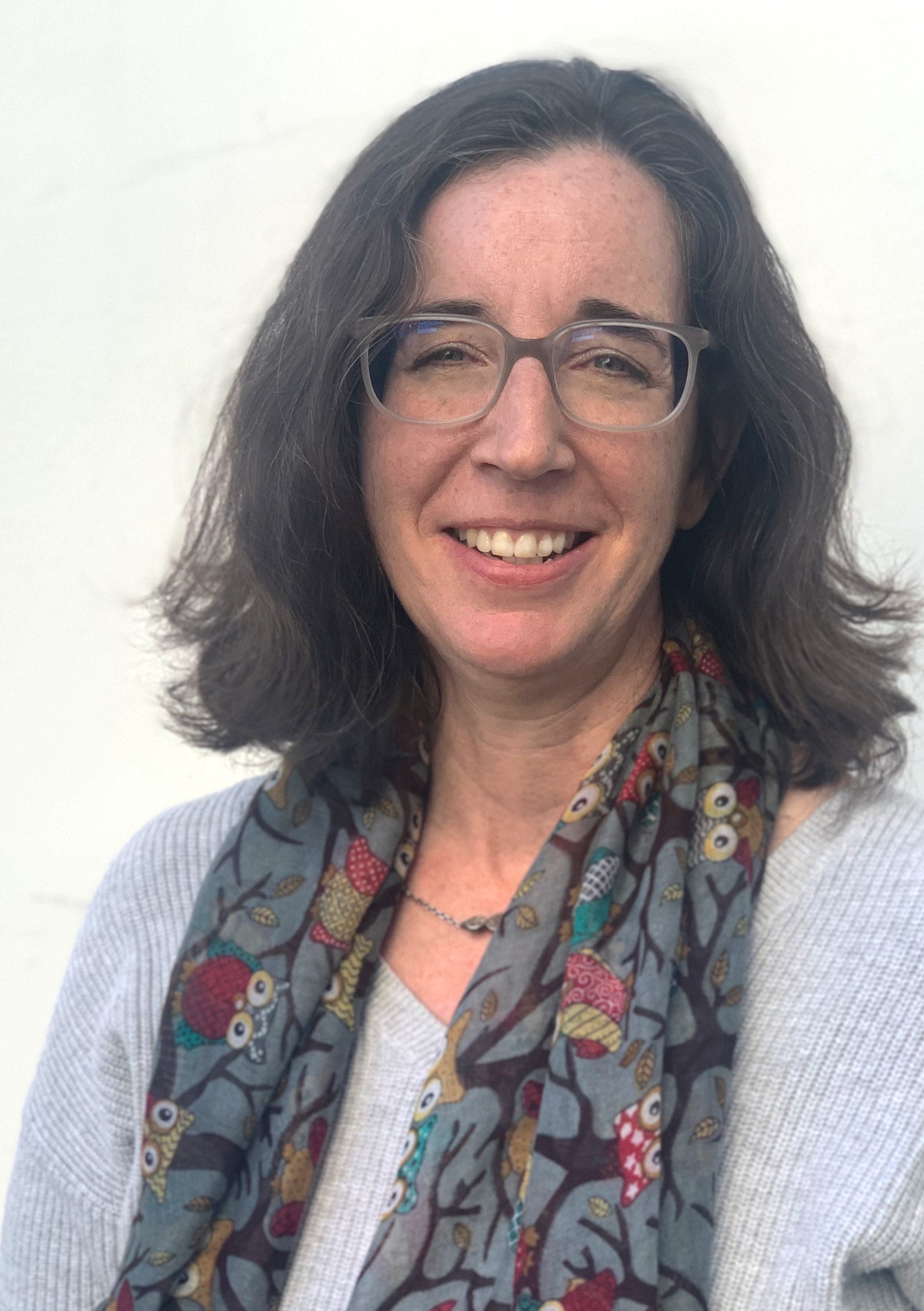 Tilla Edmunds - Woman with shoulder length long brown hair, glasses, and a smile.  She is wearing a light blue v-neck sweater and a grey patterned scarf around her neck.