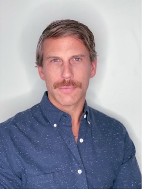 Man wearing a denim blue collared shirt with sandy colored hair and a mustache.