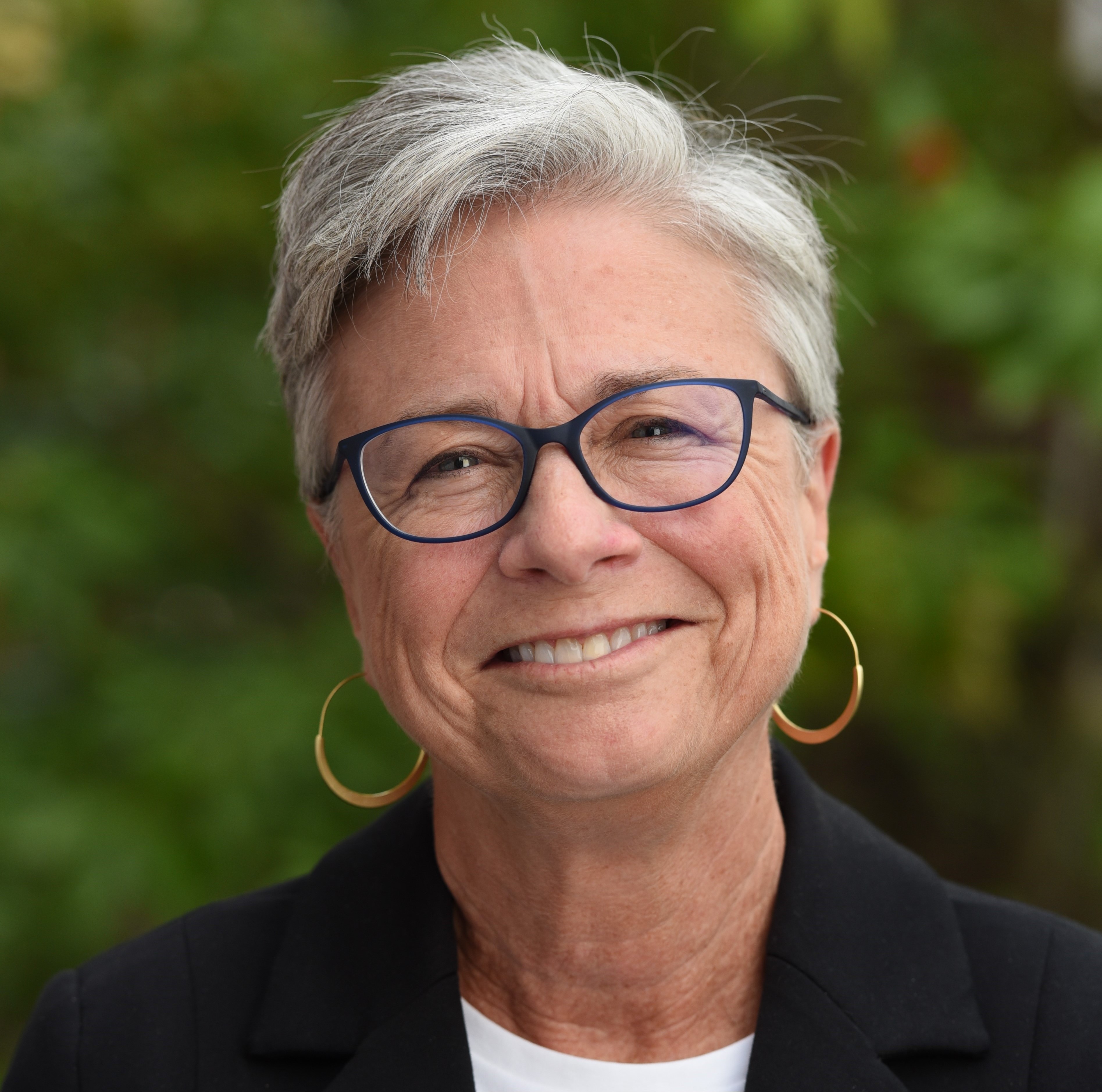 Smiling caucasian woman with short white hair, dark framed glasses, wearing gold hoop earrings and a black blazer.