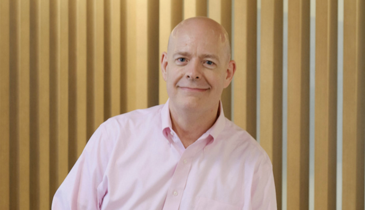 Caucasian man with shaved head, wearing a light pink collared shirt agains a background of gold vertical panels.