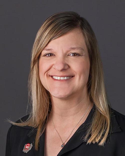 Woman with long blonde hair, wearing a black blouse and a lapel pin