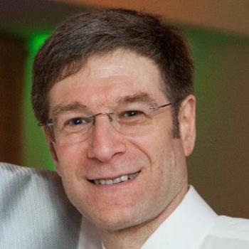 Man smiling with brown hair wearing glasses and a white collared shirt.
