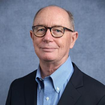 Smiling caucasian man with glasses, wearing a light blue collared shirt and black blazer.