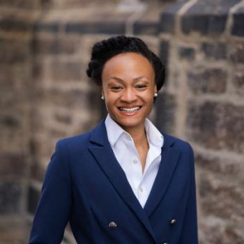 Professional portrait of a smiling woman wearing a navy blazer and white shirt, standing in front of a stone wall.