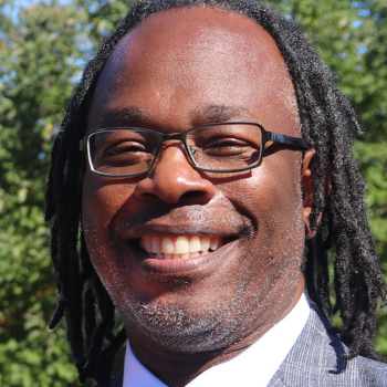 African American man smiling with short deadlocks, a slat and pepper beard, and dark framed glasses.