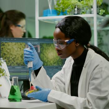 An African American scientist wearing a white lab coat, safety goggles, and blue gloves uses a micropipette to transfer liquid into a petri dish at a laboratory workstation.