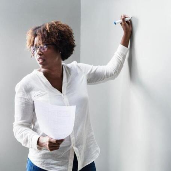 African American woman wearing glasses and a long-sleeved shite collared shirt. She is holding a piece a of paper and a dry erase marker preparing to write on a whiteboard.