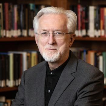 Headshot of a gray-haired man with glasses and a beard, wearing a dark suit jacket and black shirt.