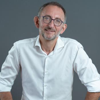 Man smiling with salt and pepper hair and beard, wearing glasses and a white collared shirt.